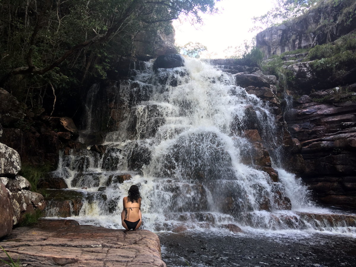 picture of waterfall cascading over rocks, with person in bikini kneeling in front away from camera.